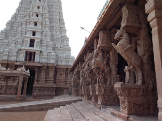 A grand temple structure showcasing intricate stone carvings and sculptures. The temple features a large towering gopuram with detailed designs, and a side view of a colonnade with horse statues on pillars. The architecture is majestic and ancient, indicative of a significant cultural and historical site.