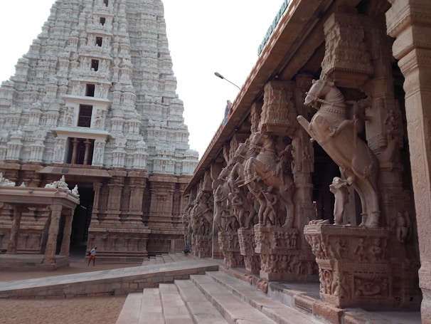 A grand temple structure showcasing intricate stone carvings and sculptures. The temple features a large towering gopuram with detailed designs, and a side view of a colonnade with horse statues on pillars. The architecture is majestic and ancient, indicative of a significant cultural and historical site.