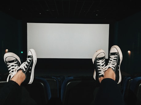 A dimly lit cinema hall with two pairs of feet wearing black and white sneakers propped up on the back of theater seats. The screen is blank, and the ambiance suggests a casual, laid-back setting.