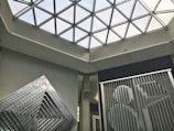 A modern architectural interior featuring a geometric glass ceiling with triangular panels. Below the ceiling, abstract art installations with linear patterns and bold designs are displayed against neutral colored walls, creating a contemporary and artistic ambiance.
