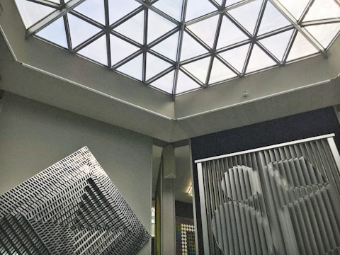A modern architectural interior featuring a geometric glass ceiling with triangular panels. Below the ceiling, abstract art installations with linear patterns and bold designs are displayed against neutral colored walls, creating a contemporary and artistic ambiance.