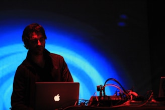 A person is operating a laptop with cables and electronic equipment on a table in front of them. The background is illuminated with vibrant blue concentric circles, creating a dynamic and energetic atmosphere. The scene is dimly lit, emphasizing the contrast between the subject and the vivid background.