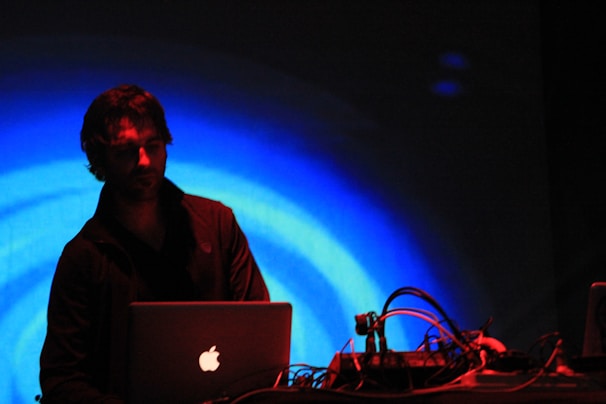 A person is operating a laptop with cables and electronic equipment on a table in front of them. The background is illuminated with vibrant blue concentric circles, creating a dynamic and energetic atmosphere. The scene is dimly lit, emphasizing the contrast between the subject and the vivid background.