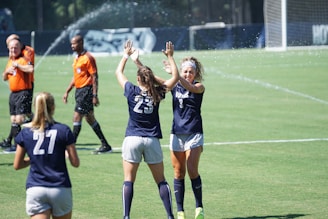 Two female soccer players wearing navy blue uniforms are high-fiving each other on a soccer field. One wears a headband and the numbers 3 and 23 are visible on their shirts. Another player with number 27 on her shirt stands nearby. In the background, referees in orange shirts walk across the field, and water sprinklers can be seen in action.