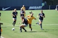 A group of female soccer players are actively competing during a soccer match. One player in a yellow jersey is attempting to take control of the ball, surrounded by several players in navy blue jerseys. The background shows a sports field with a sign for the Big East Conference.