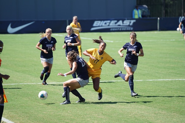 A group of female soccer players are actively competing during a soccer match. One player in a yellow jersey is attempting to take control of the ball, surrounded by several players in navy blue jerseys. The background shows a sports field with a sign for the Big East Conference.