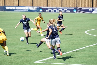 A group of female soccer players in action on the field, with some wearing navy blue uniforms and others in yellow. The scene depicts an intense game moment as players are focused on getting the ball. The grass is vibrant green, and there is a banner in the background indicating past achievements.