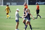 Several women are on a soccer field during a game. Two players in navy blue uniforms are exchanging a handshake with a person in a cap and athletic attire. An additional player in a yellow uniform is visible in the background, along with referees dressed in orange shirts.
