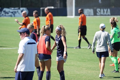 A group of female soccer players in uniforms are gathered on a grass field. In the background, referees in orange shirts and black shorts are visible. Some players are engaged in conversation or huddling together. A goal post can be seen in the distance.