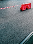 A red plastic barrier is positioned on a paved asphalt surface with a red and white striped tape extending from it. The scene is minimalistic, focusing on the textures of the rough pavement and the bright color of the barrier.
