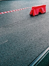 A red plastic barrier is positioned on a paved asphalt surface with a red and white striped tape extending from it. The scene is minimalistic, focusing on the textures of the rough pavement and the bright color of the barrier.