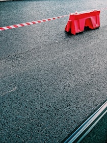 A red plastic barrier is positioned on a paved asphalt surface with a red and white striped tape extending from it. The scene is minimalistic, focusing on the textures of the rough pavement and the bright color of the barrier.