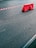 A red plastic barrier is positioned on a paved asphalt surface with a red and white striped tape extending from it. The scene is minimalistic, focusing on the textures of the rough pavement and the bright color of the barrier.