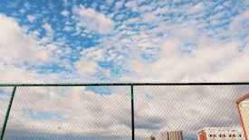 A chain-link fence stretches across the foreground with a background of a partly cloudy sky. The sky features a mix of fluffy white clouds scattered across a vibrant blue backdrop. Below, there's a partial view of several buildings and structures.