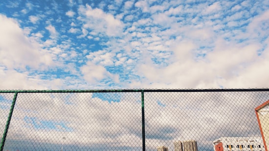 A chain-link fence stretches across the foreground with a background of a partly cloudy sky. The sky features a mix of fluffy white clouds scattered across a vibrant blue backdrop. Below, there's a partial view of several buildings and structures.