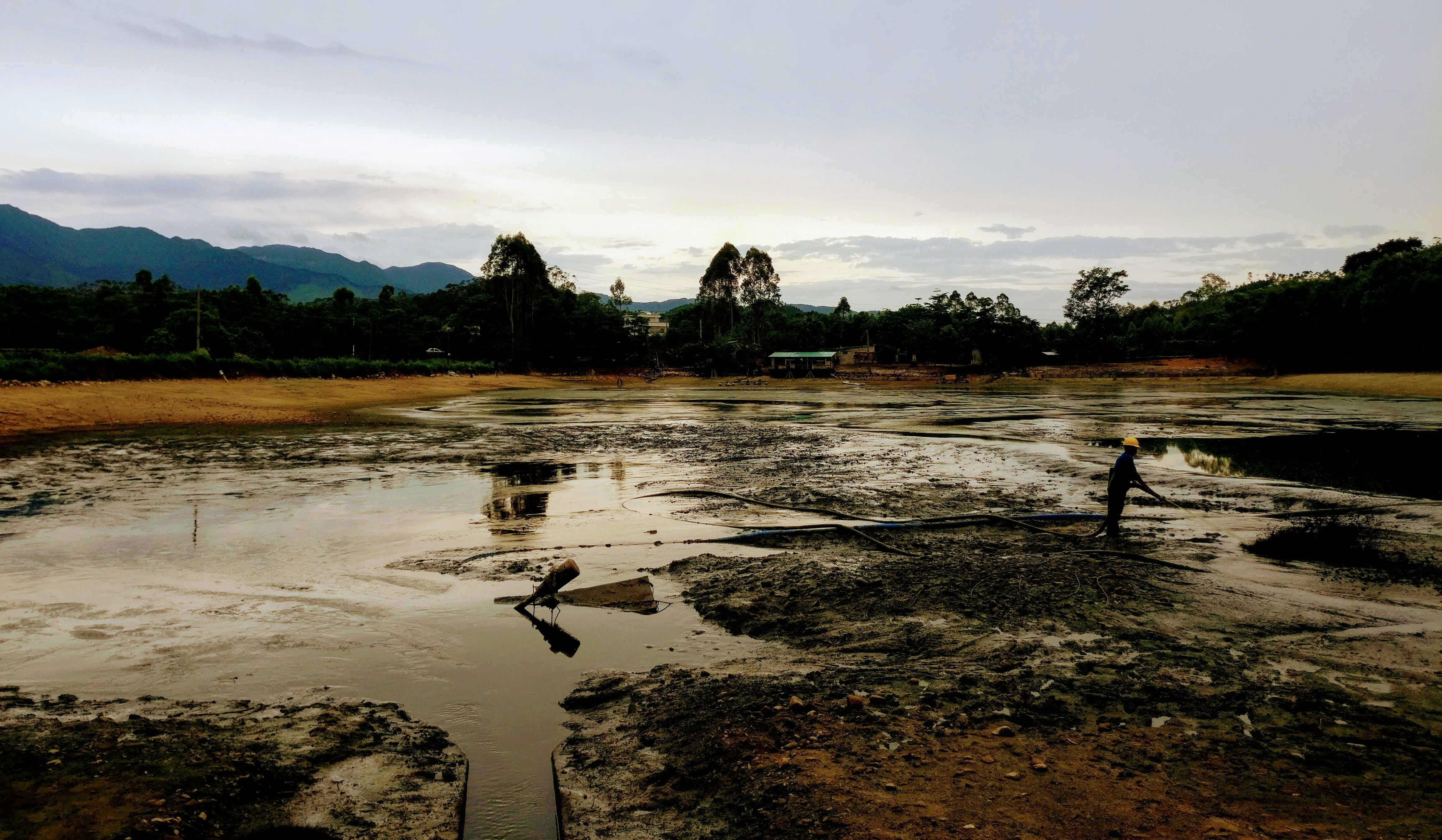 A person wearing a yellow safety helmet is standing on a muddy, barren landscape with large hoses visible on the ground. In the background, dense greenery and trees border the area, and mountains are silhouetted against a cloudy sky.