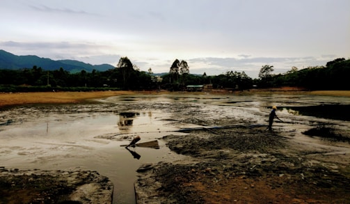 A person wearing a yellow safety helmet is standing on a muddy, barren landscape with large hoses visible on the ground. In the background, dense greenery and trees border the area, and mountains are silhouetted against a cloudy sky.