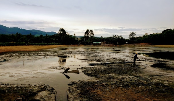 A person wearing a yellow safety helmet is standing on a muddy, barren landscape with large hoses visible on the ground. In the background, dense greenery and trees border the area, and mountains are silhouetted against a cloudy sky.