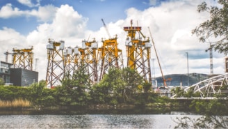 Several large, yellow industrial structures stand tall against a partly cloudy sky. These structures are surrounded by cranes and are situated near a body of water with trees and greenery in the foreground. In the background, there are distant hills and a few modern buildings, along with a white bridge to the right.