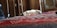 A cat lies on its back on a colorful patterned bedspread. The room features wooden furniture, including wardrobes and a chair in the corner.