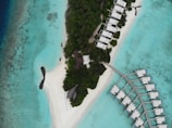 Aerial view of a tropical island with dense green foliage and white sandy beaches surrounded by crystal clear turquoise water. There are several bungalows or villas spread across the island, some on the shore and others on stilts over the water, connected by a wooden pathway. Beach chairs and umbrellas are arranged on the sandy areas.