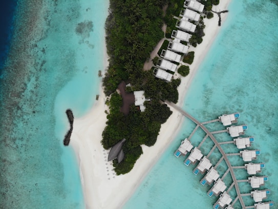 Aerial view of a tropical island with dense green foliage and white sandy beaches surrounded by crystal clear turquoise water. There are several bungalows or villas spread across the island, some on the shore and others on stilts over the water, connected by a wooden pathway. Beach chairs and umbrellas are arranged on the sandy areas.