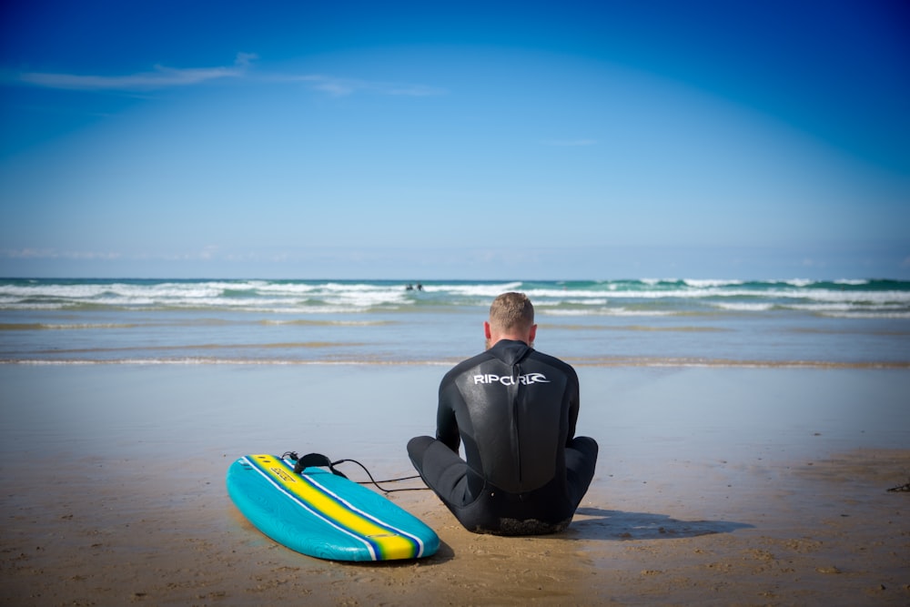 surfer sits on sand beside surfboard at the beach