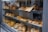A bakery display window showcasing a variety of baked goods, including loaves of bread, pastries, muffins, and croissants. The items are neatly arranged on multiple shelves, some labeled with a paper sign. The reflection of people can be seen on the glass, indicating a busy street outside.