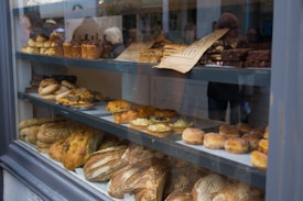 A bakery display window showcasing a variety of baked goods, including loaves of bread, pastries, muffins, and croissants. The items are neatly arranged on multiple shelves, some labeled with a paper sign. The reflection of people can be seen on the glass, indicating a busy street outside.