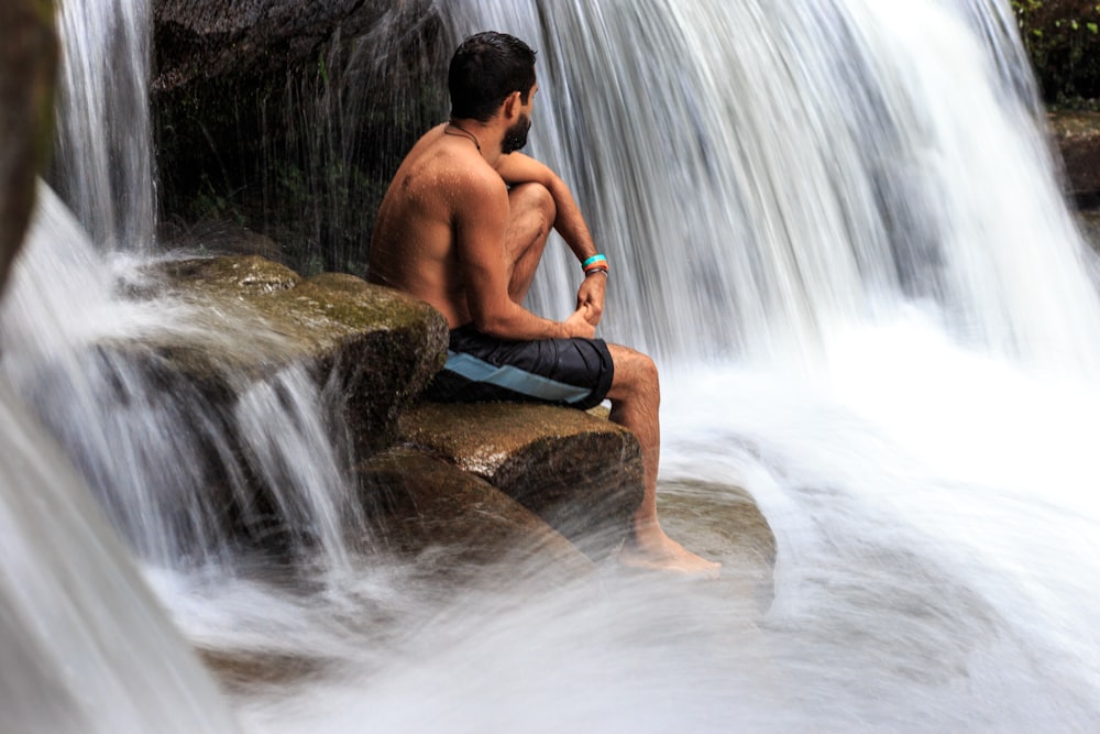 topless man sits on rock between waterfall