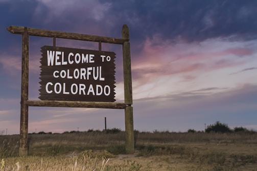 A rustic wooden sign stands in an open field, displaying the words 'Welcome to Colorful Colorado' against a backdrop of a dramatic evening sky with shades of purple, pink, and blue. The landscape is grassy with some bushes and power lines visible in the distance.
