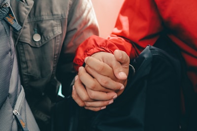 Two people holding hands, one wearing a red jacket and the other wearing a green jacket with a button. The hands are entwined, showing a connection or support between them.