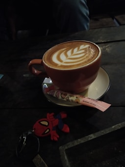 A cup of coffee with latte art in the shape of a leaf rests on a saucer, accompanied by a wrapped wafer stick with cute cartoon designs. Nearby, a keychain featuring a small superhero figure is visible on the dark wooden table.