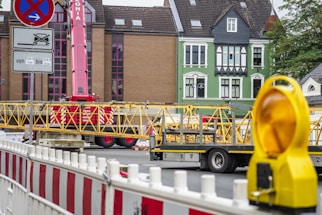 A construction site with a red crane and yellow structural elements on a truck. There are buildings in the background with distinct architecture, including a green façade. Barricades and a yellow warning light are visible in the foreground.