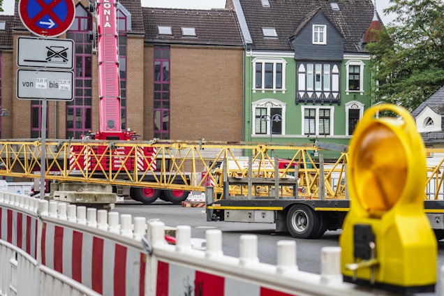 A construction site with a red crane and yellow structural elements on a truck. There are buildings in the background with distinct architecture, including a green façade. Barricades and a yellow warning light are visible in the foreground.
