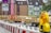 A construction site with a red crane and yellow structural elements on a truck. There are buildings in the background with distinct architecture, including a green façade. Barricades and a yellow warning light are visible in the foreground.