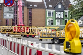 A construction site with a red crane and yellow structural elements on a truck. There are buildings in the background with distinct architecture, including a green façade. Barricades and a yellow warning light are visible in the foreground.