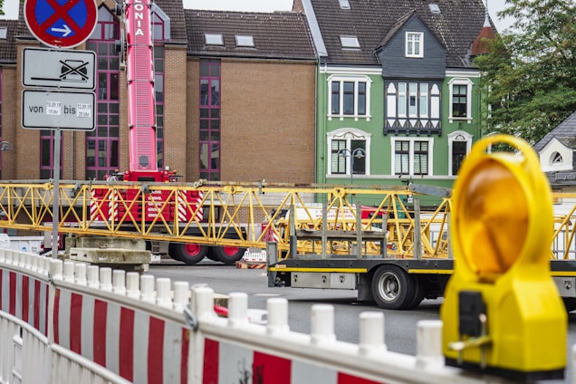 A construction site with a red crane and yellow structural elements on a truck. There are buildings in the background with distinct architecture, including a green fa&ccedil;ade. Barricades and a yellow warning light are visible in the foreground.