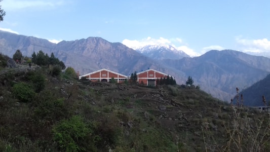 Two red-roofed buildings are nestled in a lush, green landscape with terraced vegetation. Majestic mountains with snow-capped peaks provide a dramatic backdrop under a blue sky with scattered clouds.