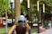 A person with long hair wearing a white and blue baseball cap with the text 'LA GUERRILLA' is standing in a park. The background features tall, candle-like street lamps, trees, a palm tree, and a few benches.