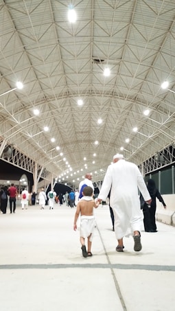Several people are walking under a large, illuminated structure with a high ceiling. The walkway is spacious and well-lit by numerous overhead lights. The focus is on an adult and a child holding hands, both wearing traditional white garments commonly associated with pilgrimage.