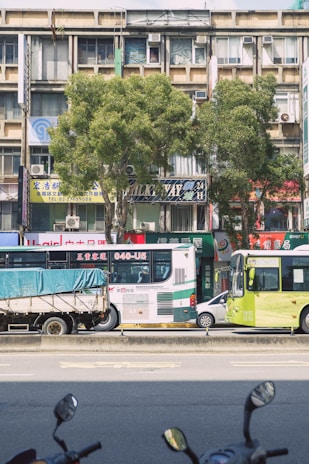 A busy urban street scene with vehicles parked on the road, including two buses and a truck with a blue tarp. Behind the vehicles, there's a row of mid-rise buildings with various signs in multiple languages, trees lining the street, and visible air conditioning units attached to the buildings.