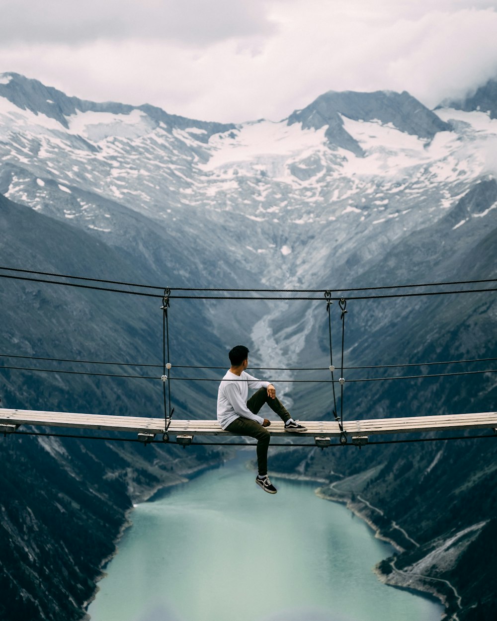 man wearing white crew-neck long-sleeved shirt sitting on beige wooden footbridge viewing body of water and mountain under white skies during daytime