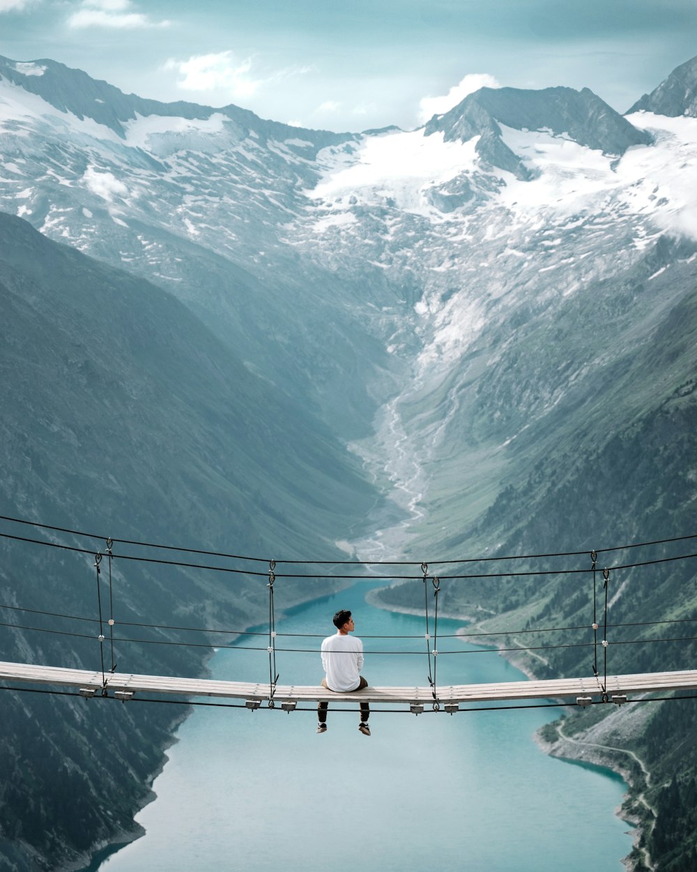 man wearing white shirt siting on bridge overlooking at mountain