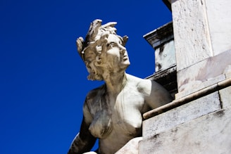 A stone statue depicting a mythological figure is captured against a deep blue sky. The figure has a detailed, weathered appearance, suggesting age and exposure to the elements. The statue features intricate carvings, including serpentine elements in the hair, and is part of a larger architectural structure.