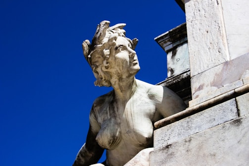 A stone statue depicting a mythological figure is captured against a deep blue sky. The figure has a detailed, weathered appearance, suggesting age and exposure to the elements. The statue features intricate carvings, including serpentine elements in the hair, and is part of a larger architectural structure.