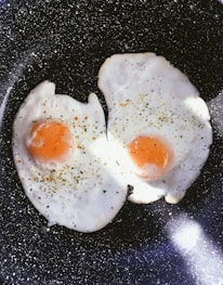 Two sunny-side-up eggs are being cooked in a black, speckled frying pan. The eggs are sprinkled with herbs and seasoning, creating a visually appealing contrast against the pristine white of the egg whites and the vibrant orange of the yolks. The lighting highlights the texture of the eggs and adds a warmth to the composition.