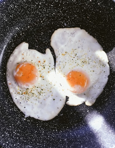 Two sunny-side-up eggs are being cooked in a black, speckled frying pan. The eggs are sprinkled with herbs and seasoning, creating a visually appealing contrast against the pristine white of the egg whites and the vibrant orange of the yolks. The lighting highlights the texture of the eggs and adds a warmth to the composition.