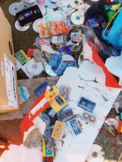A collection of old and discarded media items including several cassette tapes, CDs, and a VHS tape scattered on the ground. There's also some cardboard packaging, torn paper, and a variety of litter among the items, suggesting a disorganized and neglected area.