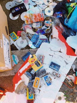 A collection of old and discarded media items including several cassette tapes, CDs, and a VHS tape scattered on the ground. There's also some cardboard packaging, torn paper, and a variety of litter among the items, suggesting a disorganized and neglected area.